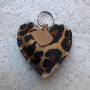 May include: Heart-shaped keychain with a leopard print design. The keychain features a metal ring and a small leather tag with the initials "MO". The edges are stitched with white thread.