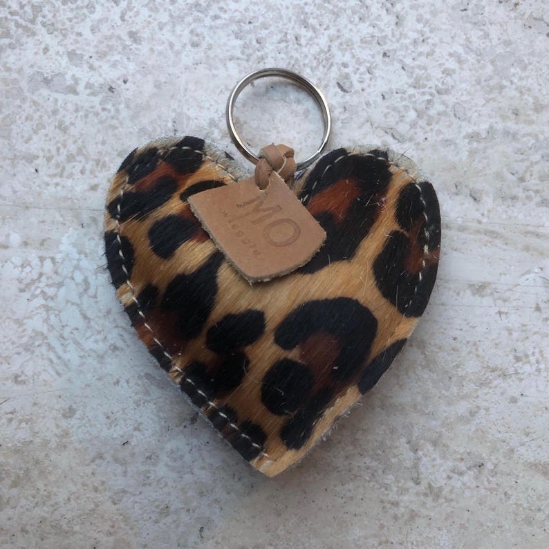May include: Heart-shaped keychain with a leopard print design. The keychain features a metal ring and a small leather tag with the initials "MO". The edges are stitched with white thread.