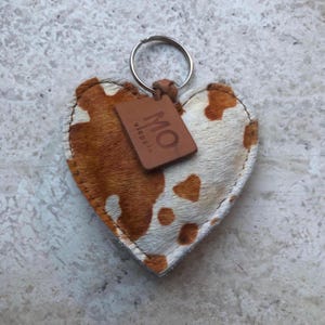 May include: Heart-shaped keychain with a cowhide pattern in brown and white. A small, brown leather tag with the letters "MO" is attached. The keychain has a silver metal ring.