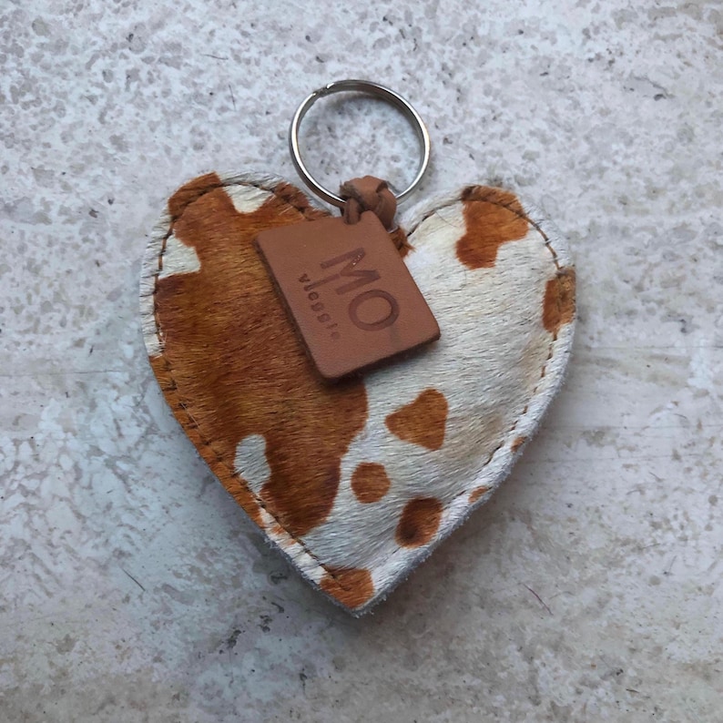 May include: Heart-shaped keychain with a cowhide pattern in brown and white. A small, brown leather tag with the letters "MO" is attached. The keychain has a silver metal ring.