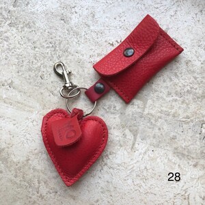 May include: A red leather keychain featuring a heart-shaped charm and a small rectangular pouch with a snap closure. The heart charm has a small leather tag attached with the embossed text MO SARAH. A silver metal lobster clasp is attached to the ring.