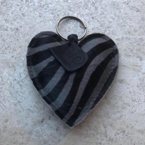 May include: Heart-shaped keychain with a zebra stripe pattern in black and gray. The keychain has a silver metal ring and a small black tag attached. The tag has the letters "MO" on it.