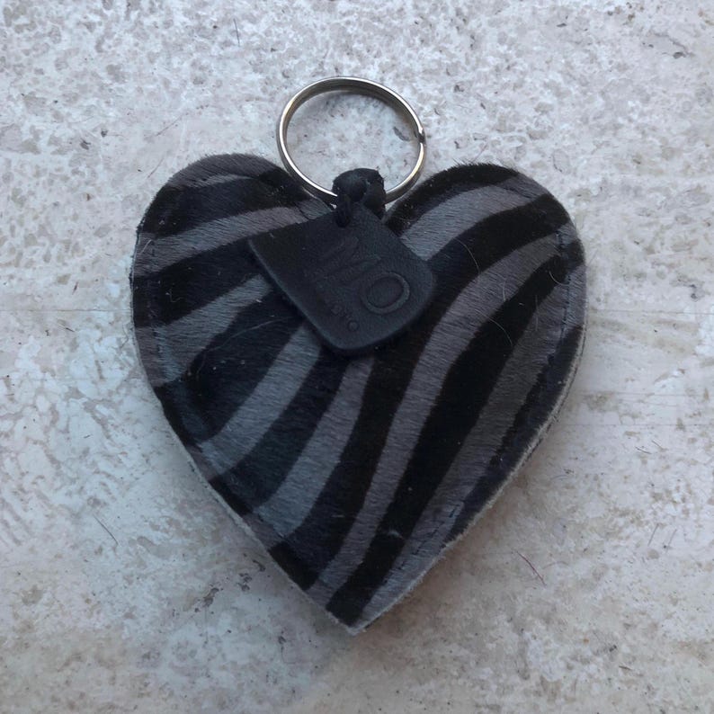 May include: Heart-shaped keychain with a zebra stripe pattern in black and gray. The keychain has a silver metal ring and a small black tag attached. The tag has the letters "MO" on it.