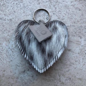 May include: Heart-shaped keychain with a cowhide pattern in black and white. The keychain has a silver ring and a small, rectangular tag with the word "virgoic" printed on it. The edges of the heart are stitched with white thread.