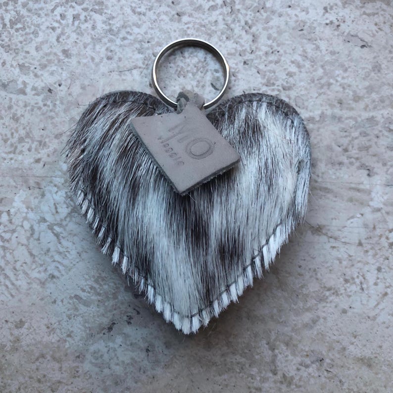 May include: Heart-shaped keychain with a cowhide pattern in black and white. The keychain has a silver ring and a small, rectangular tag with the word "virgoic" printed on it. The edges of the heart are stitched with white thread.