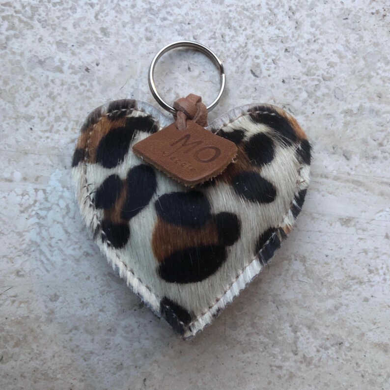 May include: Heart-shaped keychain with a cowhide pattern in black, brown, and white. The keychain has a silver ring and a small, brown leather tag with the letters "MO". The edges are stitched with white thread.