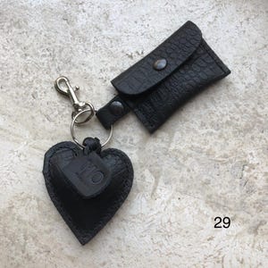 May include: A black keychain featuring a heart-shaped charm with a crocodile-embossed texture and a small rectangular pouch with a snap closure. The heart charm has a small leather tag embossed with the letters MO. The number 29 is visible.