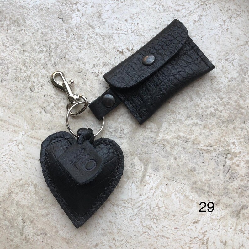 May include: A black keychain featuring a heart-shaped charm with a crocodile-embossed texture and a small rectangular pouch with a snap closure. The heart charm has a small leather tag embossed with the letters MO. The number 29 is visible.