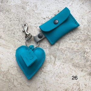 May include: A bright turquoise leather keychain featuring a heart-shaped charm with the embossed letters MO and a small rectangular pouch attached to a silver metal lobster clasp and split ring. The number 26 is printed in the bottom right corner.