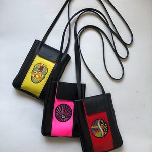 Può includere: Tre borse a tracolla in pelle nera con disegni ricamati colorati. Le borse hanno una chiusura a lembo e una lunga tracolla regolabile. I disegni includono un teschio giallo, un sole rosa e un fungo rosso.