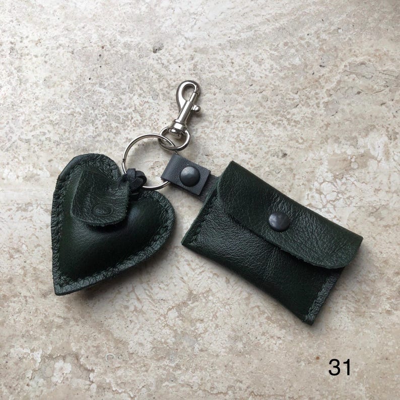 May include: A dark green leather keychain featuring a heart-shaped charm and a small rectangular pouch with a snap closure, both attached to a silver-toned metal split ring and a lobster clasp. The number 31 is printed in the bottom right corner.