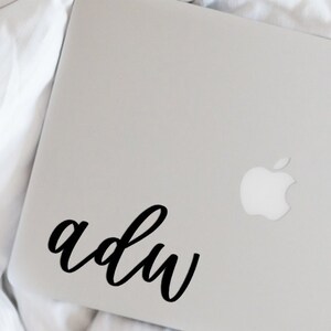 May include: A silver laptop with a black cursive sticker that says "a d u w" on the lid. The Apple logo is visible on the lid.