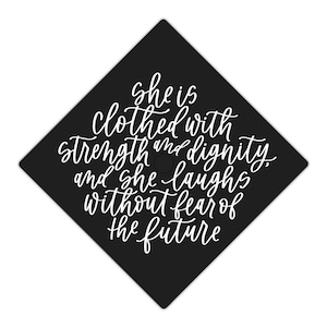 以下が含まれることがあります： 白いスクリプト文字が入った黒い卒業キャップ。テキストは「彼女は強さと尊厳に包まれ、未来を恐れずに笑う」と書かれています。