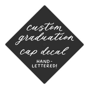 Könnte beinhalten: Ein schwarzer Abschlusskappen-Aufkleber mit weißem, kursivem Text, der "custom graduation cap decal" und "HAND-LETTERED!" lautet. Der rautenförmige Aufkleber ist für die Anbringung auf einer Abschlusskappe konzipiert.