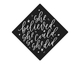 May include: Black graduation cap with white text that reads "She believed she could, so she did."  The text is written in a cursive font and is surrounded by small white stars.