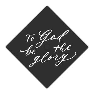May include: A black graduation cap with the phrase "To God be the glory" in white script. The cap is square-shaped and set against a white background. The text is centered and in a flowing, elegant font.