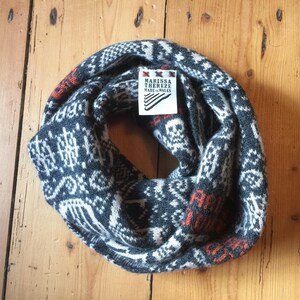 May include: A gray, white, and orange knit infinity scarf with a geometric pattern. The scarf has a tag that reads "MARISSA THEREZE MADE IN WALES". The scarf is displayed on a wooden surface.