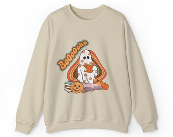 Puede incluir: Una sudadera beige con un gráfico de un fantasma leyendo un libro con el texto "BOO000ks" en una fuente retro. El fantasma está sentado en una pila de libros con una calabaza y otras decoraciones de otoño.