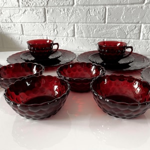 May include: A set of ruby red glass dishes, including teacups, saucers, plates, and bowls. The glassware has a textured, bubble-like pattern. The dishes are arranged on a white surface, with a white brick wall in the background.