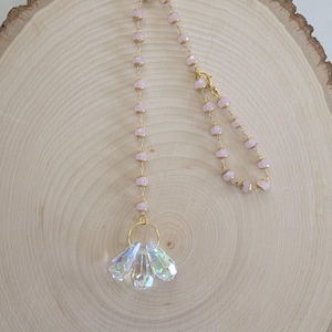 May include: A delicate, handmade crystal suncatcher with a pink beaded chain and gold-colored accents. The suncatcher features a cluster of iridescent, faceted crystals suspended from a gold ring. The beaded chain is composed of small, light pink beads.