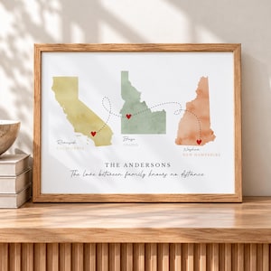 May include: A framed wall art print featuring watercolor maps of California, Idaho, and New Hampshire. Red hearts mark Riverside, Boise, and Nashua, connected by dashed lines. Text reads THE ANDERSONS and The love between family knows no distance.