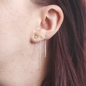 May include: Close-up of an ear with three silver earrings. One is a small stud, and the other two are dangling. One is a delicate chain, and the other is a straight bar. The earrings are simple and elegant.