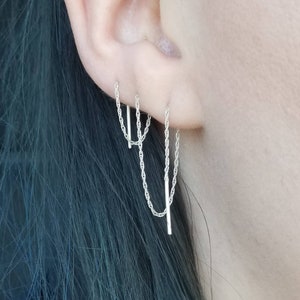 May include: Silver chain earrings with a bar and chain design. The earrings are shown on an ear, with the chain dangling down. The earrings are made of a shiny silver material.