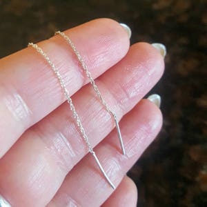 May include: A pair of silver threader earrings. Each earring has a fine chain leading to a straight bar. The earrings are shown against a skin-toned background.