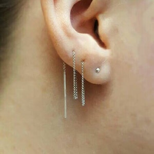 May include: Close-up of an ear with three silver earrings. One is a long, thin bar, and the other two are chain-style, dangling earrings. A small silver stud earring is also visible.