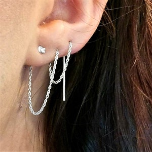 May include: Silver chain earrings with a delicate design. The earrings feature a chain that dangles from the ear, with a small stud earring visible above. The chains are connected to a small bar.