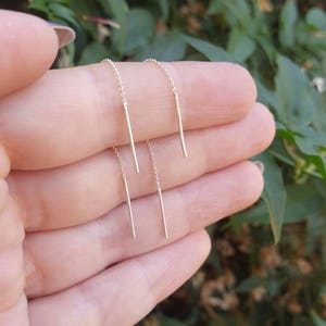 May include: Gold threader earrings. Each earring has a slender, straight bar hanging from a fine chain. The earrings are shown against a blurred green backdrop, possibly outdoors.