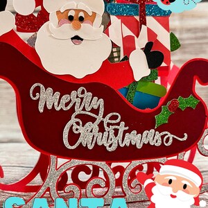 K&ouml;nnte beinhalten: Eine rote und wei&szlig;e 3D-Weihnachtskarte mit einem roten Schlitten, Geschenken und dem Schriftzug "Frohe Weihnachten".