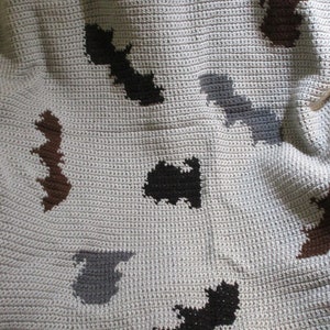 May include: A crocheted blanket with a white background and a pattern of bats in various shades of brown, black, and gray.
