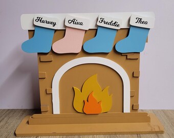 May include: A wooden fireplace with a fire and four stockings hanging above it. The stockings are decorated with the names Harvey, Aiwa, Freddie, and Theo.