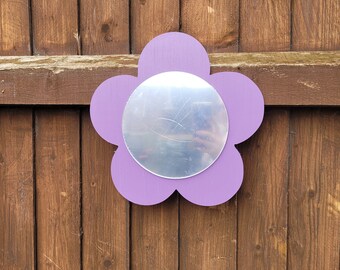 Könnte beinhalten: Ein lilafarbener, blumenförmiger Spiegel mit einer silbernen reflektierenden Oberfläche. Der Spiegel ist an einer Holzwand befestigt.