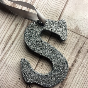 May include: A silver glitter letter S ornament with a white ribbon attached.