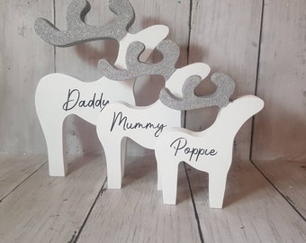 Puede incluir: Tres renos de madera blanca con cuernos de purpurina plateada. Los renos están de pie en fila, con el reno más grande en la parte posterior y el reno más pequeño en la parte delantera. Los renos están etiquetados como "Daddy", "Mummy" y "Poppie".