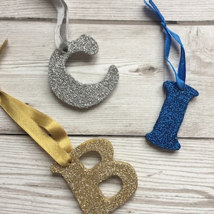May include: Three glitter letters, B, I, and C, with gold, blue, and silver glitter, respectively. Each letter is attached to a ribbon.