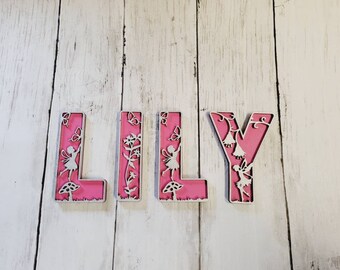 May include: Pink wooden letters spelling "LILY" with a fairy and floral design cut out. The letters are on a white wood background.