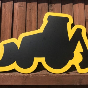 May include: A yellow silhouette of a construction vehicle, such as a bulldozer or excavator, with a black interior. The silhouette is cut out of wood and mounted on a wooden surface.