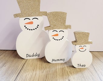 Op de afbeelding: Drie witte houten sneeuwpoppen met gouden glitterhoeden. De grootste sneeuwpop zegt "Paddy", de middelste sneeuwpop zegt "Mummy" en de kleinste sneeuwpop zegt "Theo".