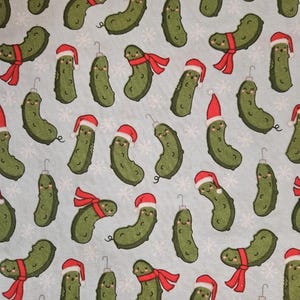 May include: A light blue fabric with a repeating pattern of cartoon pickles. Some pickles wear Santa hats, others have red scarves, and all have smiling faces. White snowflakes are scattered throughout the design.