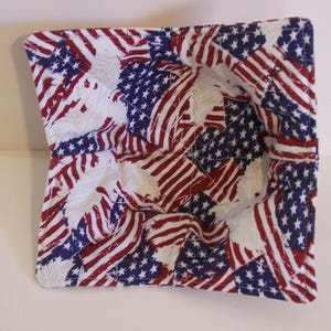 Könnte beinhalten: Ein quadratischer, gesteppter Schalenwärmer mit einem sich wiederholenden Muster der amerikanischen Flagge. Der Stoff ist weiß mit roten Streifen und blauen Sternenfeldern. Zum Halten einer Schüssel und zum Schutz der Hände vor Hitze.
