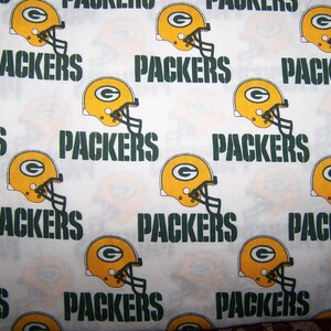 Op de afbeelding: Witte stof met een herhaald patroon van gele en groene American footballhelmen en het woord "PACKERS" in groen. De helmen hebben een groen "G"-logo.