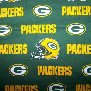Op de afbeelding: Groene stof met een herhalend patroon van het woord "PACKERS" in gele blokletters, het "G"-logo van het team en een geel en groene footballhelm. Geschikt voor knutselen of kleding.
