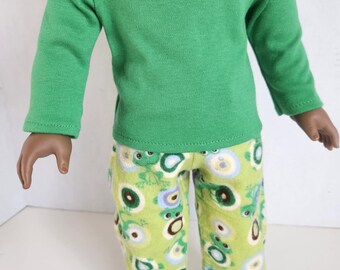 May include: A doll wearing a green long-sleeved shirt and pajama pants. The pajama pants are light green with a pattern of frogs, circles, and flowers in shades of green, blue, and white. The doll is standing.