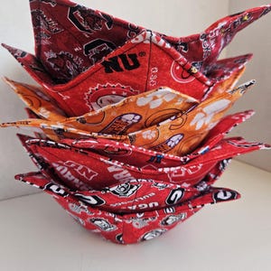 May include: A stack of four fabric bowl cozies featuring various collegiate sports team logos and mascots in red, orange, and white. The quilted bowl holders have a four-cornered, star-like shape and are designed to hold bowls securely while protecting hands from heat.