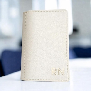 May include: A cream-colored, rectangular wallet with the initials "RN" embossed on the lower right corner. The wallet has a stitched border and a smooth, leather-like texture. The wallet is standing upright on a white surface.