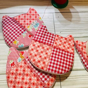 May include: Handmade cat-shaped quilted coasters in red and pink floral and gingham patterns. The coasters are made of fabric and have a stitched edge. A spool of red thread is in the background.