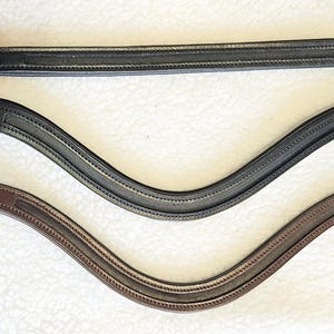 Puede incluir: Tres testeras de cuero en negro, gris y marrón. Cada testera tiene un diseño curvo con costuras decorativas y se asegura con un botón en cada extremo. Las testeras son accesorios ecuestres.